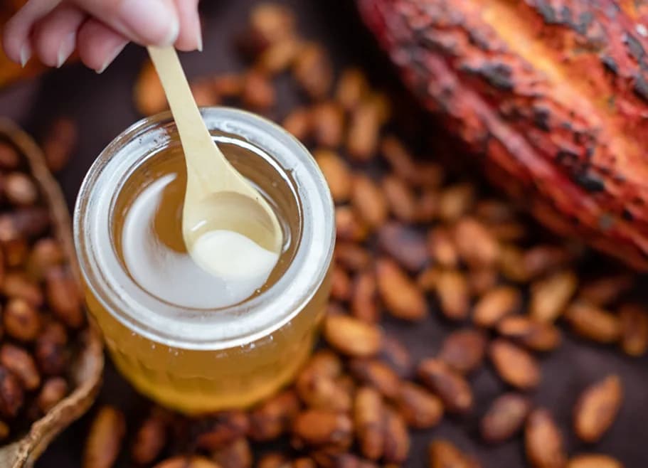 Una cuchara de madera saca miel de un frasco, sobre granos de cacao
